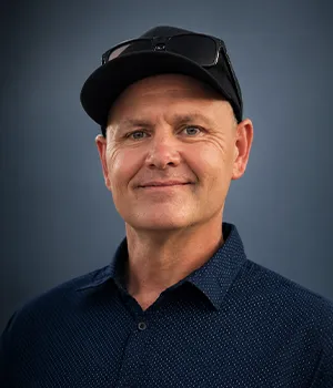 Smiling man in a black cap and sunglasses, wearing a dark blue shirt, on a blue background.