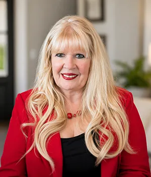 Smiling woman with long blonde hair, red blazer, and black top sitting indoors.