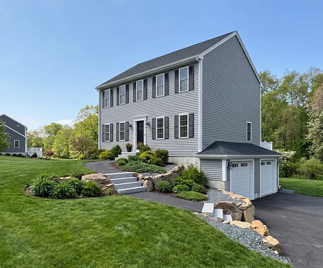 Two-story gray house with a double garage, landscaped yard, and stone steps to entrance.