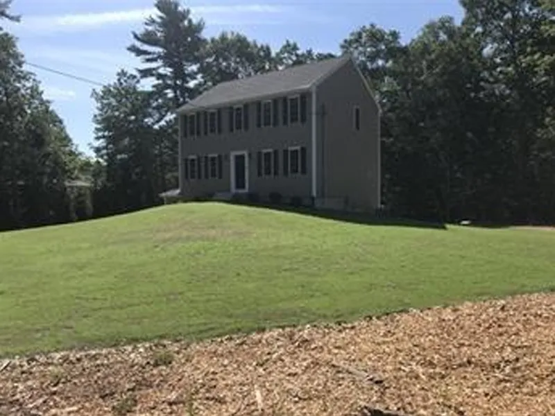 Two-story gray house on a grassy hill with trees in the background under a blue sky.