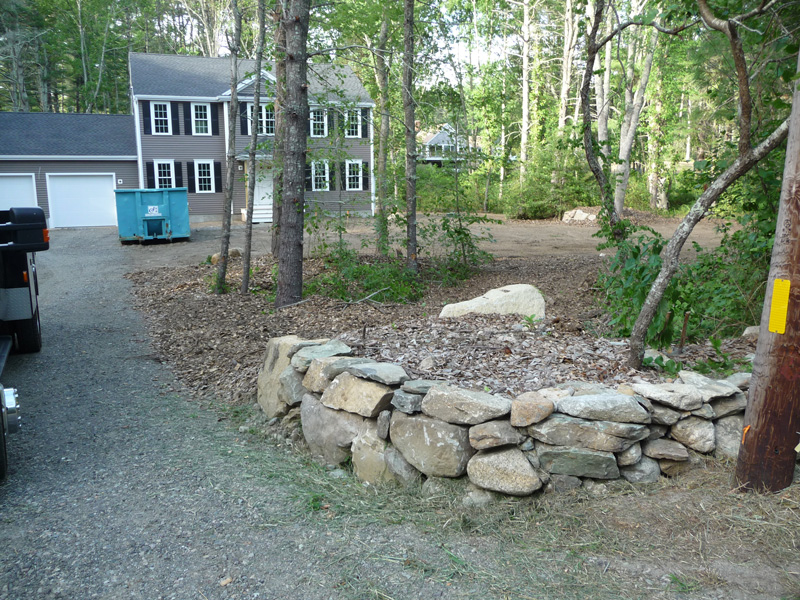 Stone wall and mulch area by driveway, with house, trees, and a blue dumpster in background.