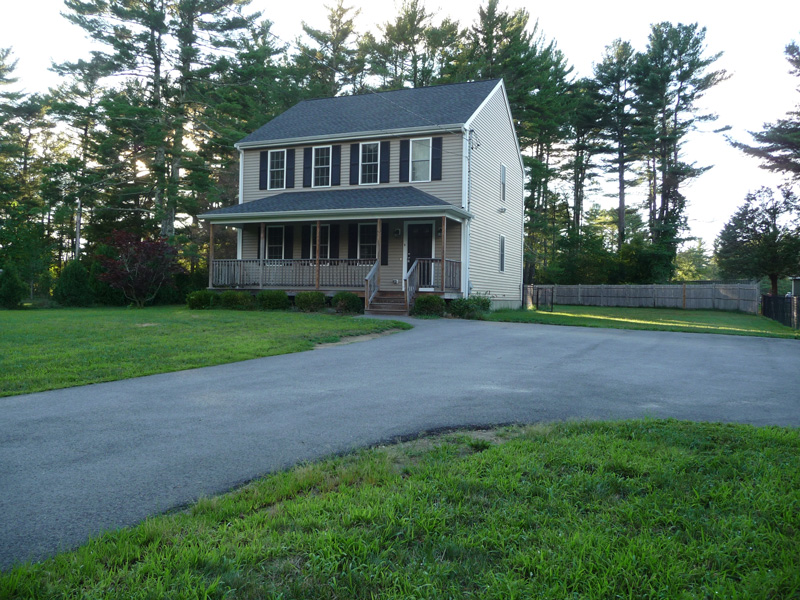 Two-story beige house with porch, large driveway, and trees in the background.