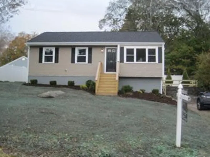 Single-story beige house with front steps, black door, and a for-sale sign on the lawn.