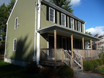 Green two-story house with a covered front porch and wooden railings on a sunny day.