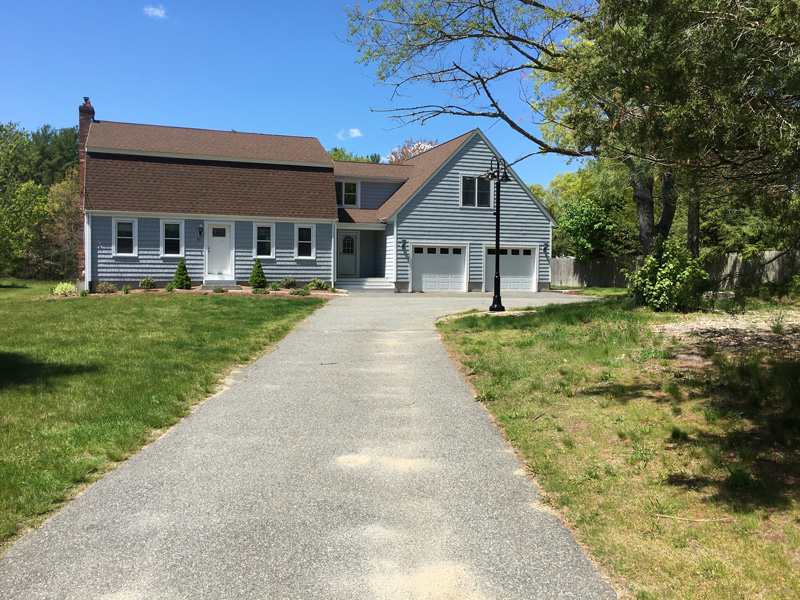 Gray two-story house with double garage and long driveway, surrounded by grass and trees.