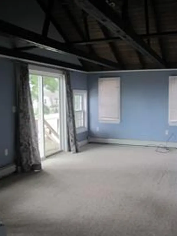 Empty blue room with carpet, exposed beams, sliding glass door, and windows with blinds.