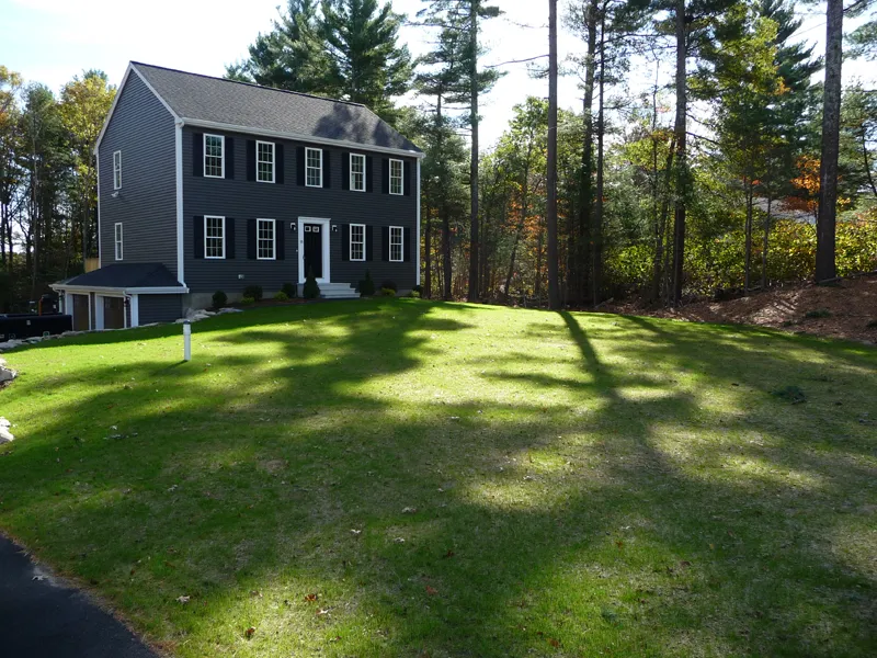 A two-story gray house with white trim sits on a large, sloped green lawn by trees.