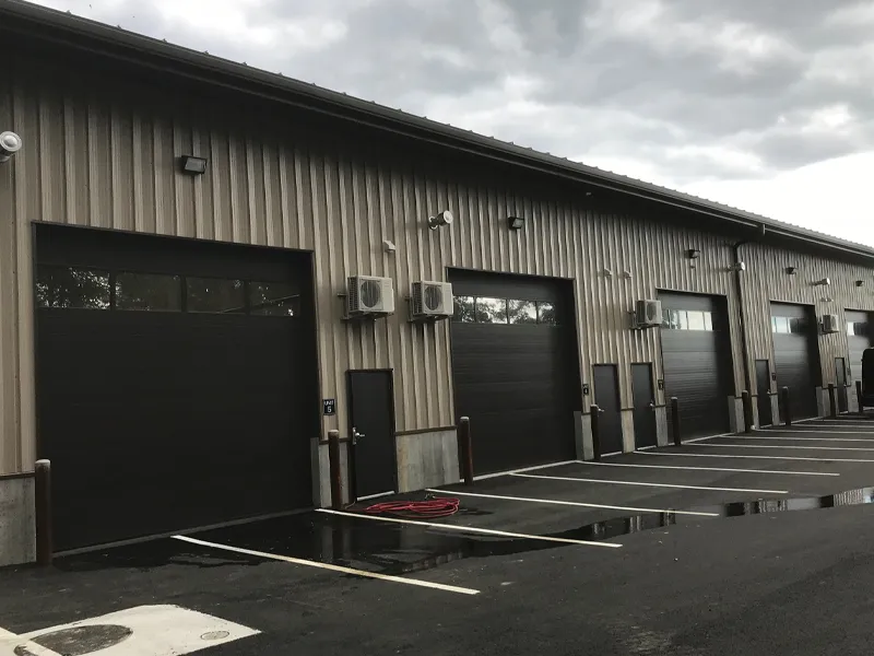 A row of closed garage doors and parking spaces outside a commercial building under cloudy skies.