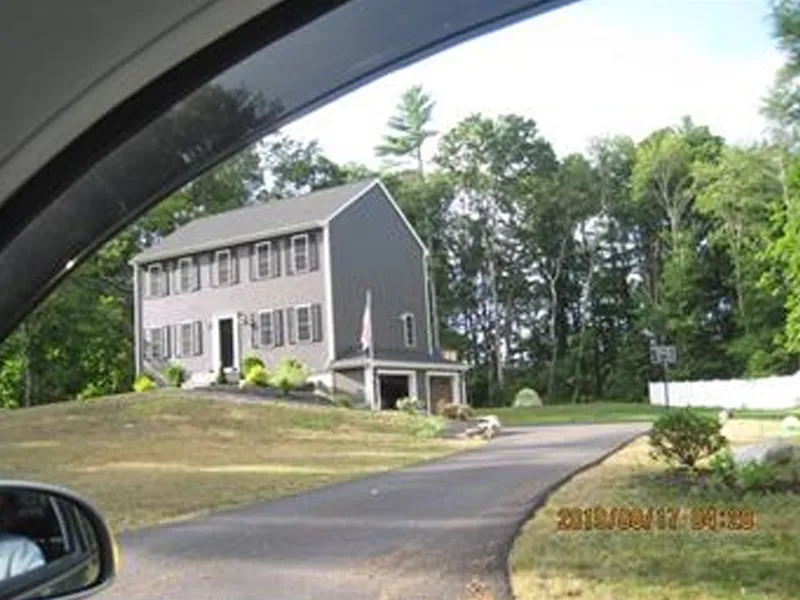 Gray two-story house with shutters, viewed from a car window, trees in the background.