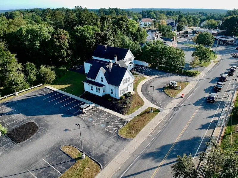 Aerial view of a white house with black roof, parking lot, and nearby streets and trees.