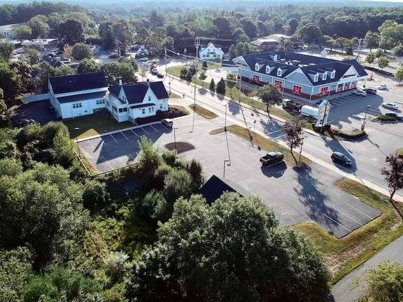 Aerial view of a suburban street with buildings, parking lots, and trees.