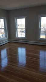 Empty room with hardwood floor, three large windows, and pale gray walls.