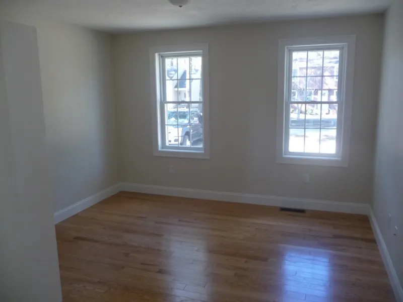 Empty room with light-colored walls, wooden floor, and two windows letting in sunlight.