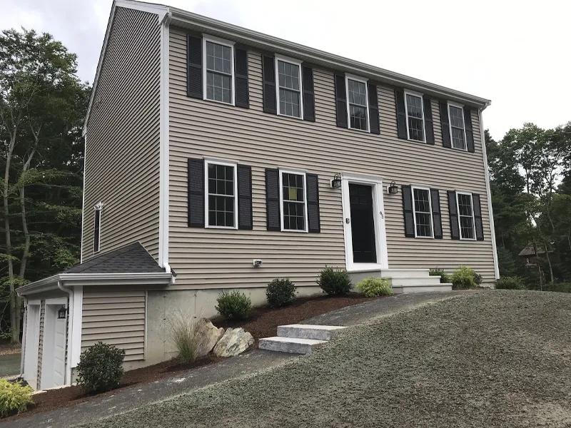 Two-story tan house with black shutters, garage, and small front steps on a sloped yard.