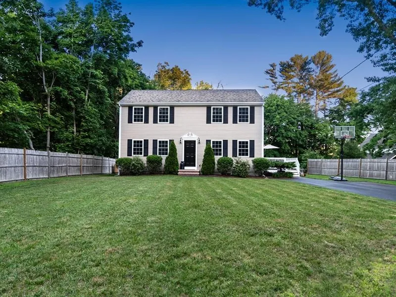 Two-story beige house with black shutters, large green lawn, and basketball hoop driveway.