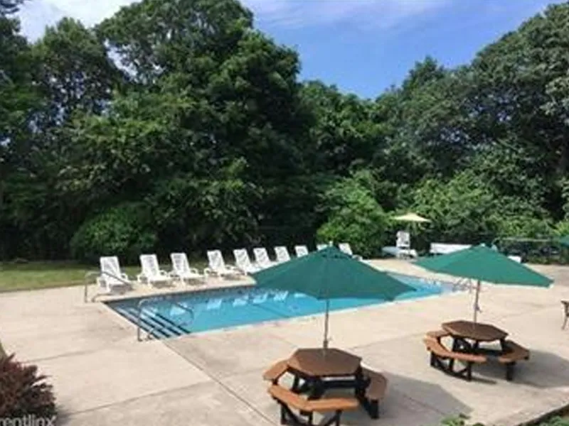 Outdoor pool with lounge chairs, picnic tables, and green umbrellas surrounded by trees.