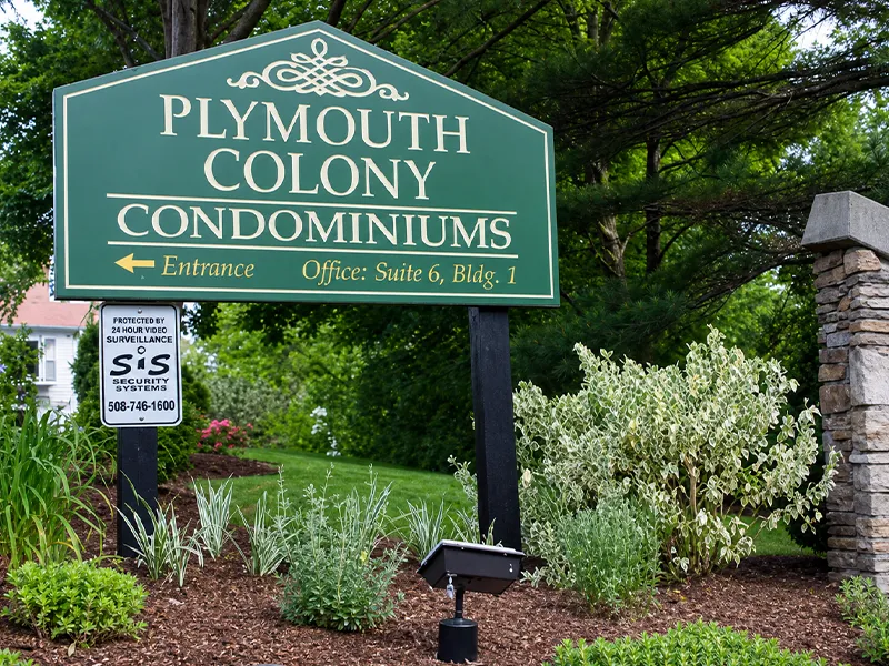 Green sign reading Plymouth Colony Condominiums by landscaped bushes and stone pillar.