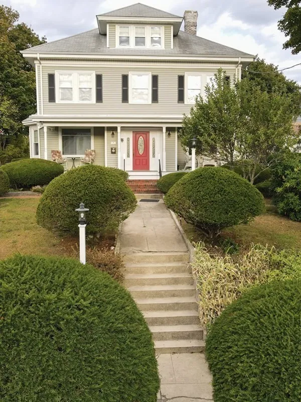 Large gray house with red door, front steps, and round, neatly trimmed green bushes.