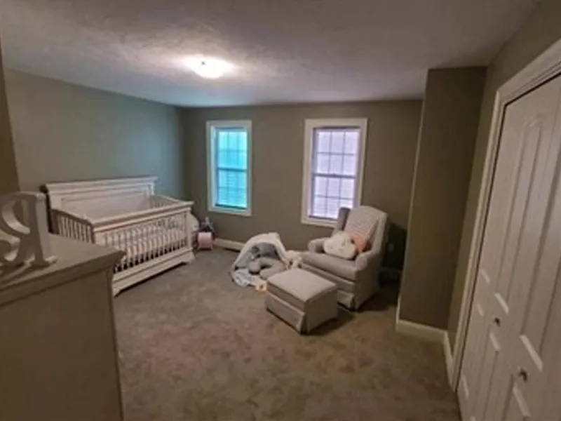 A nursery with a crib, armchair, ottoman, and two windows, in soft neutral tones.