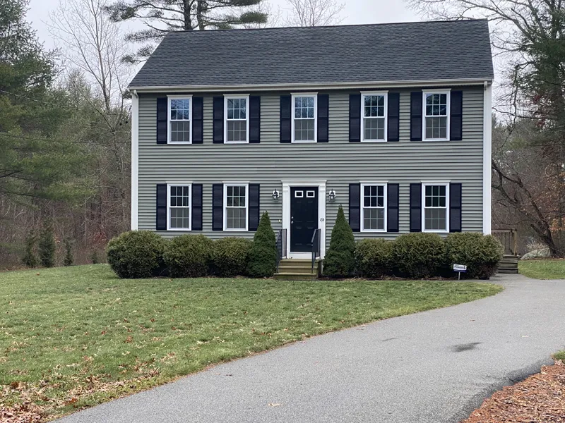 Two-story gray house with black shutters, central door, bushes, and a curved driveway.