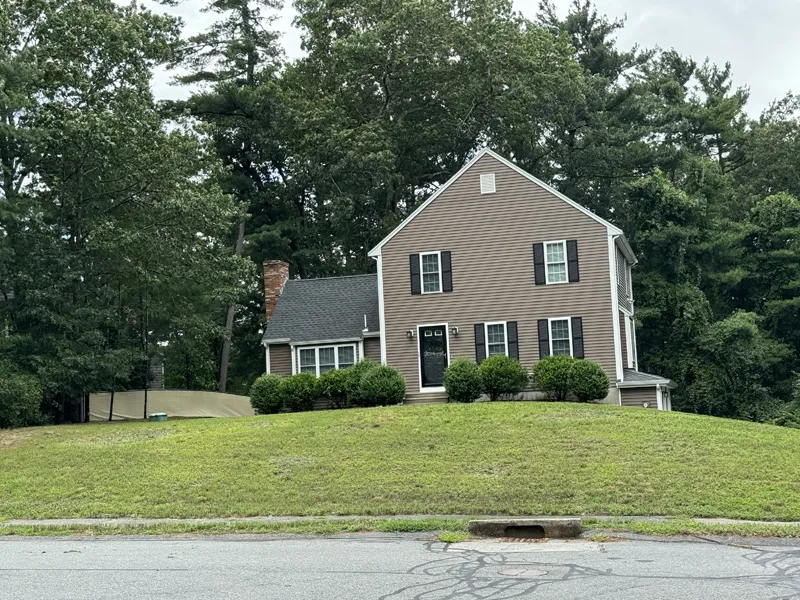 Brown two-story house with black shutters sits on a grassy hill surrounded by trees.
