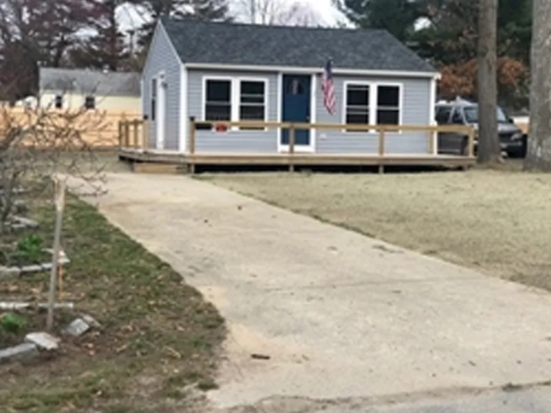 Small gray house with blue door, front porch, and American flag, viewed from driveway.