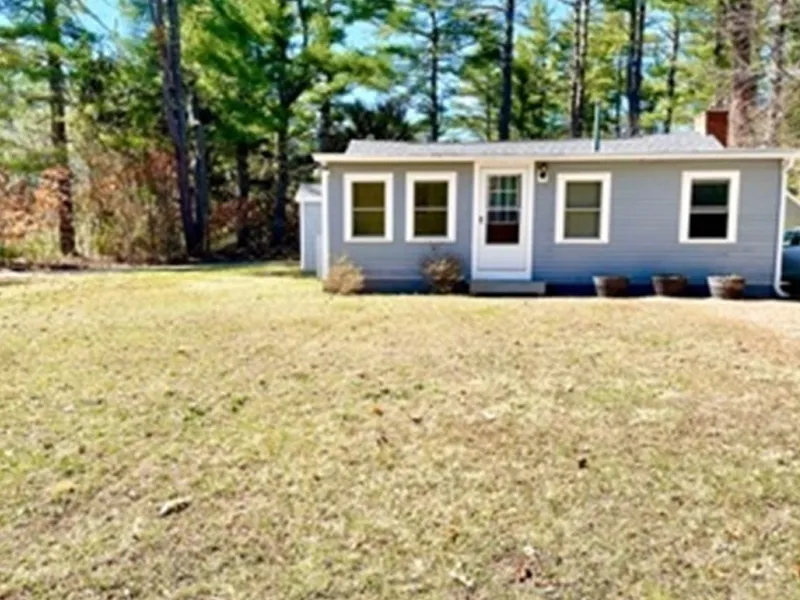 Small gray house with white trim, surrounded by grass and trees under a clear sky.