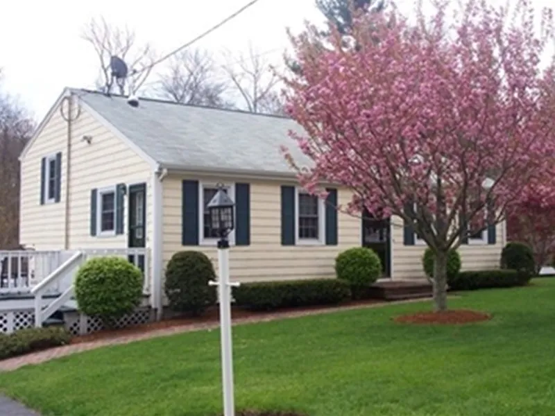 Yellow house with black shutters, a blooming pink tree, and neatly trimmed green lawn.