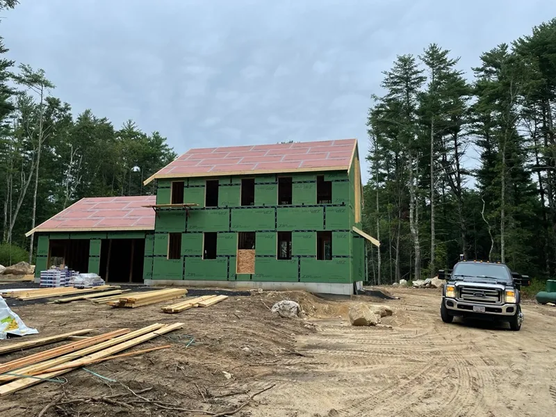 Partially constructed house with green sheathing, lumber, and a truck on a dirt lot.