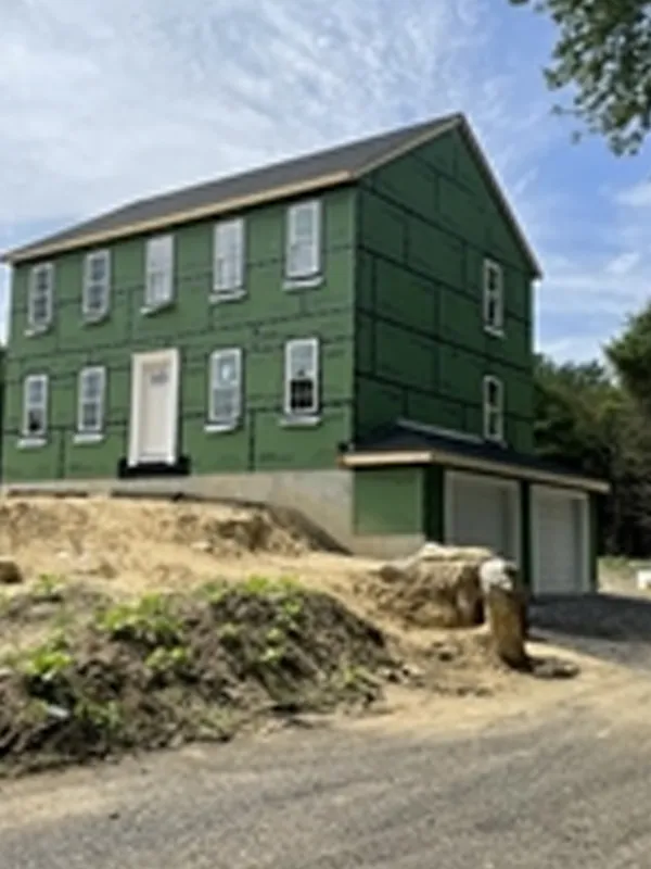 Two-story house under construction with green sheathing, next to a dirt driveway.