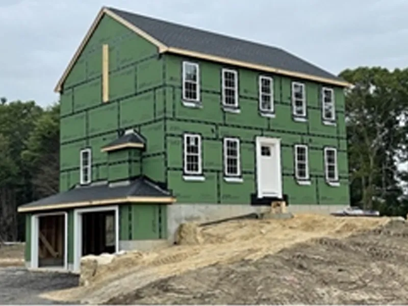 A two-story house under construction with green sheathing and an unfinished yard.