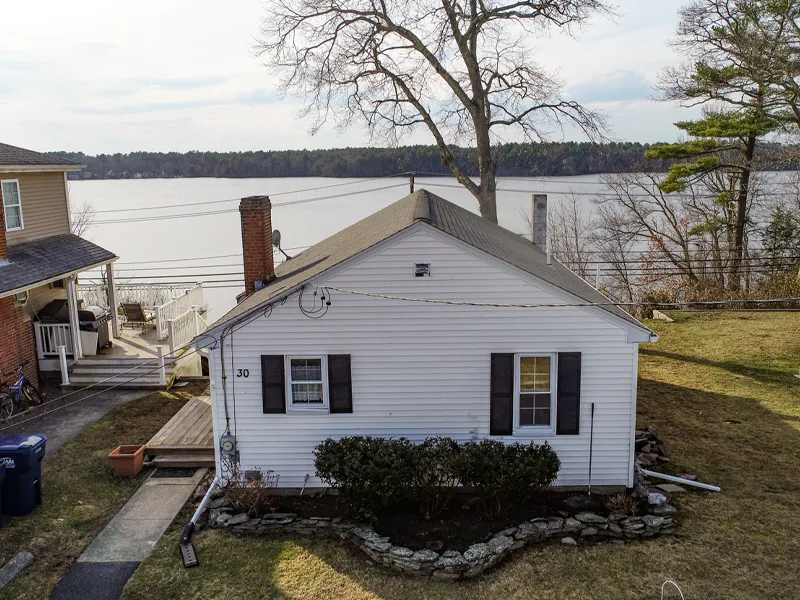 Small white house with black shutters by a lake, with trees and a stone-bordered yard.