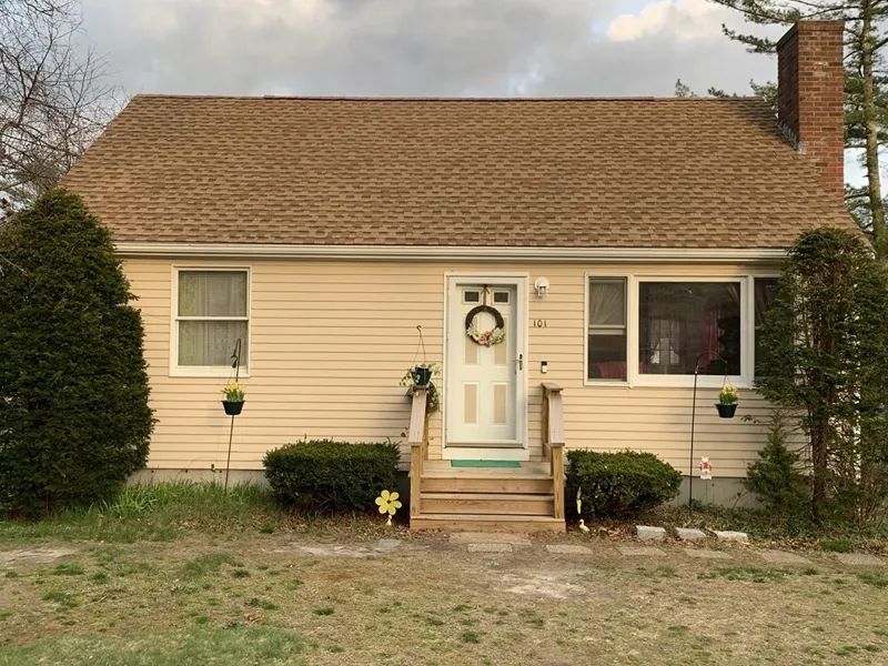 Small beige house with a porch, wreath on the door, and shrubs on either side.