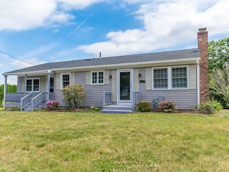 Single-story gray house with front porch, brick chimney, and a grassy lawn.