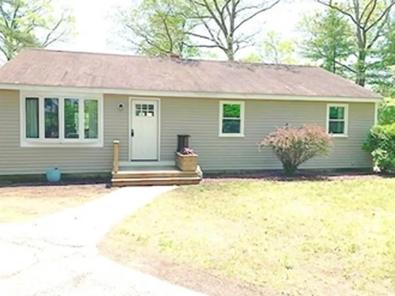Single-story beige house with white trim, front steps, small shrubs, and a grassy yard.