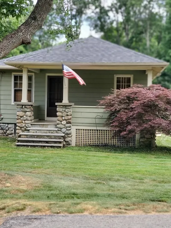 Small green house with porch, American flag, and a purple-leafed tree in front yard.