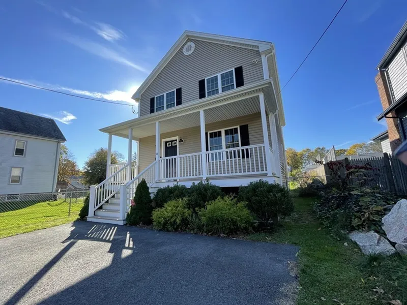 Two-story beige house with a front porch, white railing, and black shutters on a sunny day.