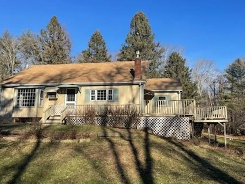 Yellow house with a deck, chimney, and large windows, surrounded by trees under blue sky.