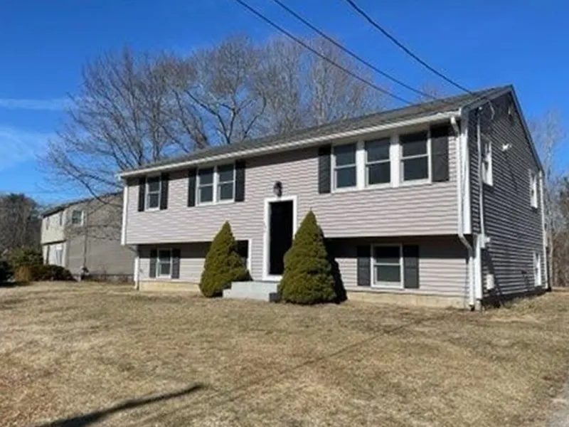 Gray split-level house with two front doors, evergreen shrubs, and a brown lawn in front.