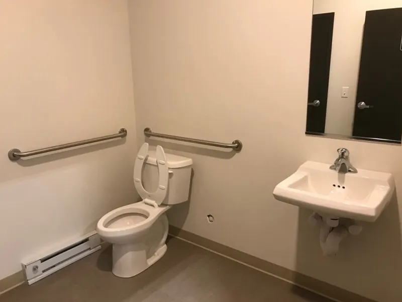 Accessible restroom with grab bars, toilet, sink, and mirror on plain beige walls.