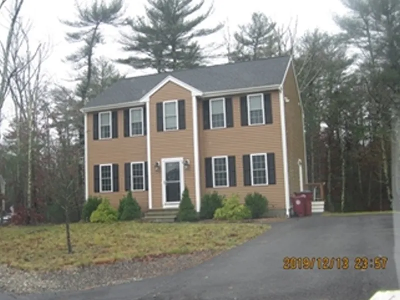 Two-story tan house with black shutters, front steps, driveway, and surrounding trees.