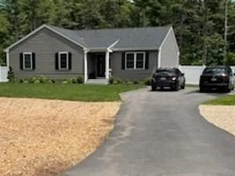 Single-story gray house with two black cars parked on a driveway, trees in the background.