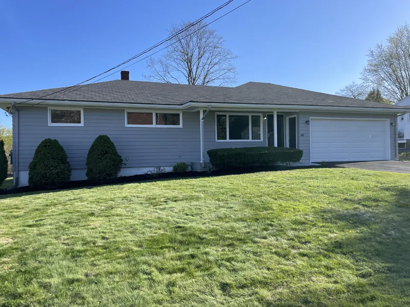 Single-story gray house with a garage, front lawn, and shrubs on a sunny day.