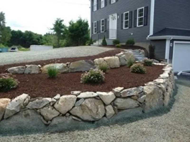 A landscaped yard with a stone retaining wall and small shrubs in front of a house.