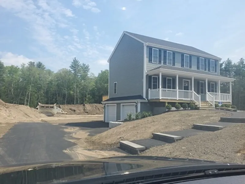 Gray two-story house with a porch, surrounded by dirt and new pavement, trees in background.
