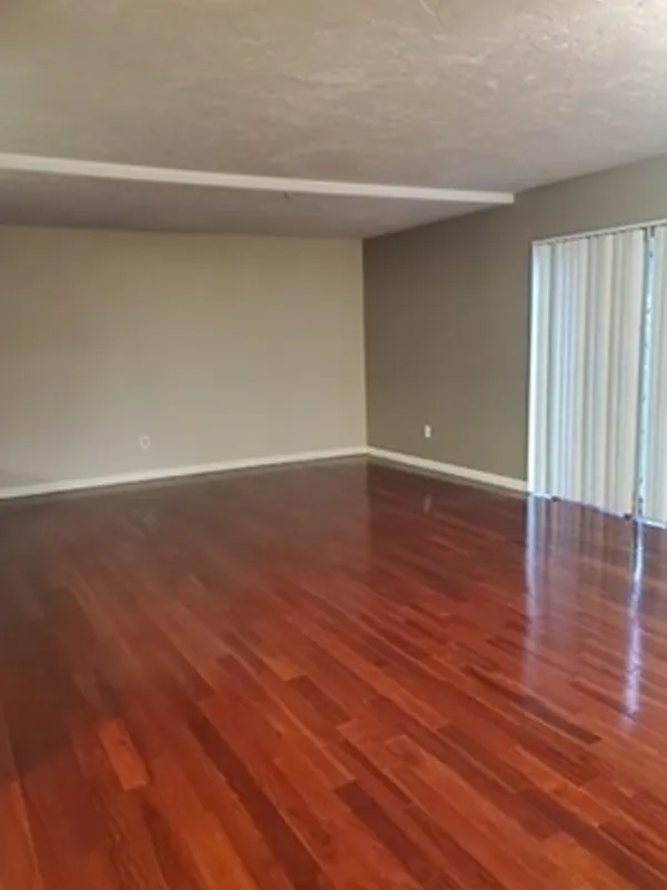 Empty room with polished hardwood floor, beige walls, and vertical blinds on sliding doors.