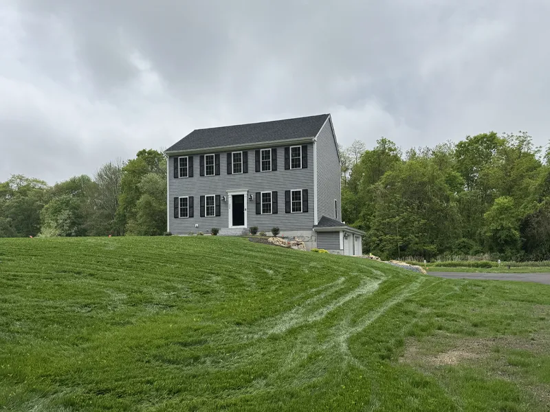 Gray two-story house with black shutters, set on a grassy hill, trees in the background.