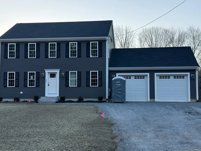 Two-story gray house with double garage and portable toilet on gravel driveway.