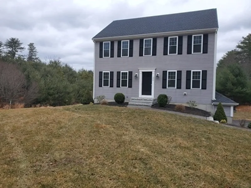 Two-story gray house with black shutters on a sloped lawn, surrounded by trees.