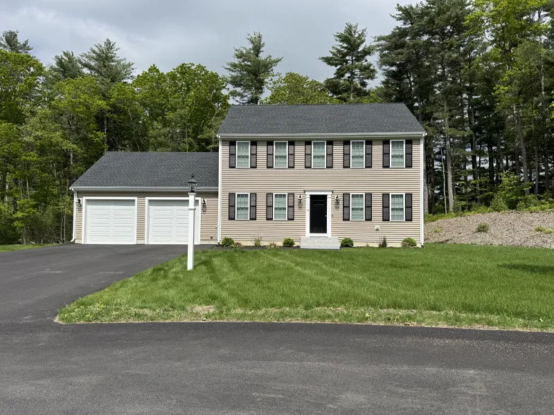 A two-story beige house with black shutters and a double garage, surrounded by trees.